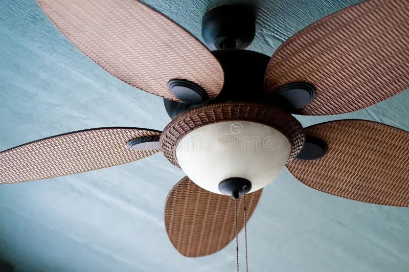 A close-up view of a ceiling fan with five textured brown woven blades and a central light fixture, mounted on a light blue ceiling.