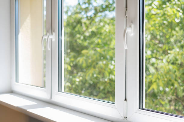 Close-up of modern white window frames with clean glass windows paneled together, showcasing bright natural light and a blurry view of green trees outside.
