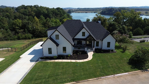 A panoramic view of a modern house showcasing complete exterior upgrades with white siding, a dark gray shingled roof, black window frames, and a curved concrete walkway next to a lake.