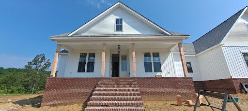 A front view of a white house under construction featuring new porch additions, including a red brick foundation, brick steps, unfinished wood support columns, and a clean gray shingled roof gable.